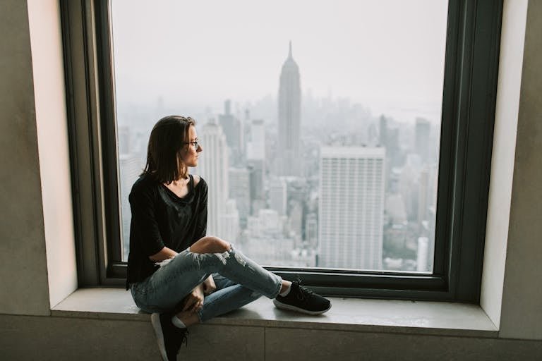 A woman sits on a windowsill, admiring the New York City skyline with the Empire State Building in view.