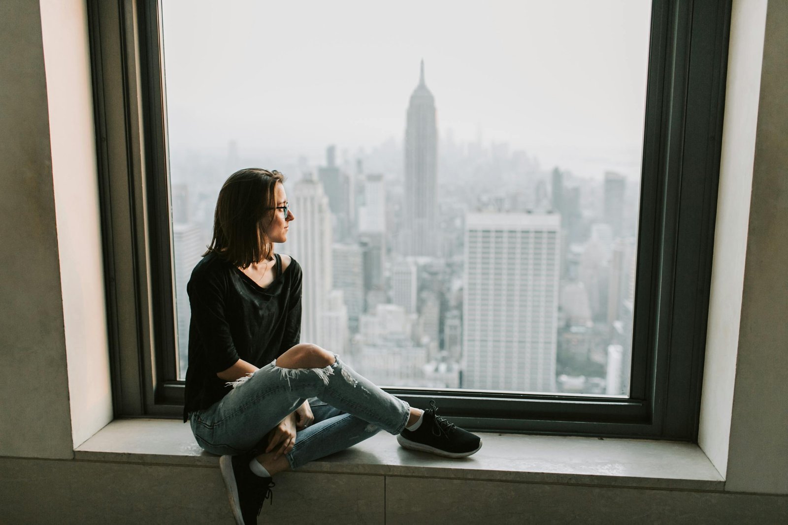 A woman sits on a windowsill, admiring the New York City skyline with the Empire State Building in view.