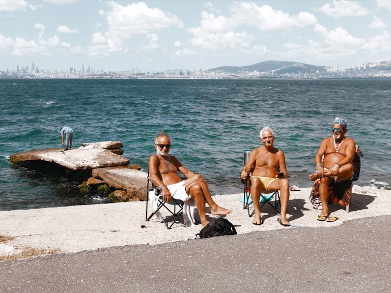 Three senior adults relaxing by the sea with a city skyline backdrop on a sunny day.