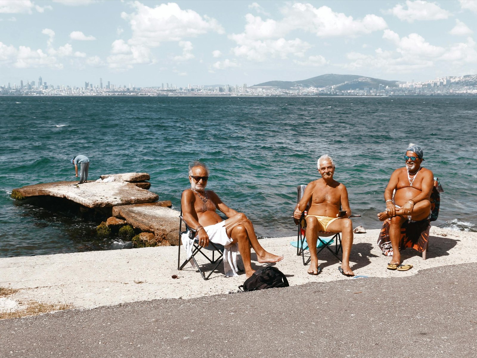 Three senior adults relaxing by the sea with a city skyline backdrop on a sunny day.